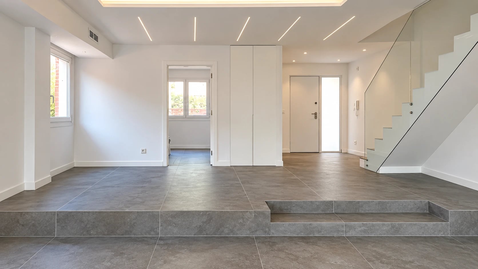 Estancia reformada con suelo gris, iluminación lineal y escalera con barandilla de vidrio