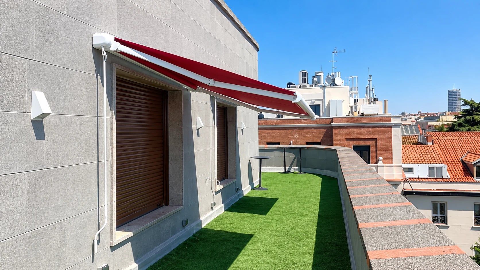 Vista de una terraza con toldo rojo, césped artificial y fachada revestida en gris