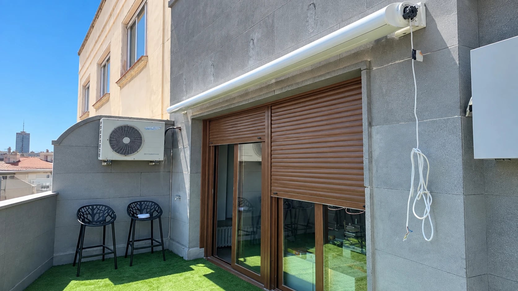 Terraza con toldo blanco recogido, persianas de madera y conexiones visibles junto a la fachada gris